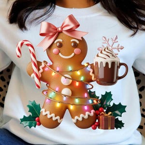 Peut inclure: Sweat-shirt blanc orné d'un bonhomme en pain d'épices. Le bonhomme en pain d'épices porte un nœud rose, des guirlandes lumineuses et tient un sucre d'orge et une tasse de chocolat chaud. Houx et un coffret cadeau complètent le look festif.