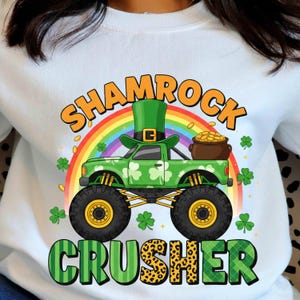 Peut inclure: Sweat-shirt blanc avec un motif de camion monstre vert orné d'un chapeau de leprechaun, de trèfles et d'un pot d'or. Le texte "SHAMROCK CRUSHER" est affiché au-dessus et en dessous du camion, sur fond d'arc-en-ciel.