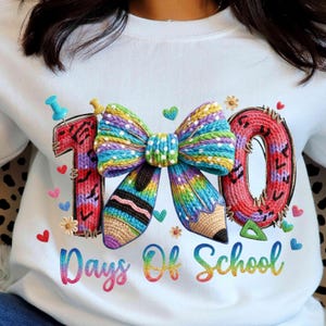 Peut inclure: Sweat-shirt blanc avec un motif coloré célébrant les 100 jours d'école. Le motif comprend un grand nœud, un crayon et les chiffres "1" et "0" dans un style tricoté, ainsi que le texte "Days Of School" aux couleurs de l'arc-en-ciel. Petits cœurs et fleurs.
