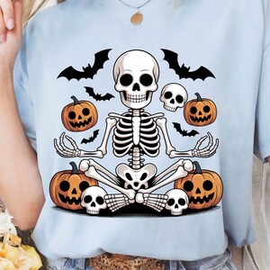 Peut inclure: T-shirt bleu clair avec un motif d'Halloween. Il présente un squelette de dessin animé en position de yoga, avec des citrouilles, des crânes et des chauves-souris. Un design amusant et effrayant pour Halloween.