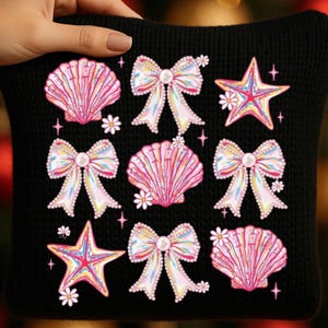 Peut inclure: Coussin noir avec un motif répété de coquillages roses, de nœuds et d'étoiles de mer. Les nœuds et les étoiles de mer ont des reflets irisés, avec de petites fleurs blanches parsemées. Le coussin est tenu par une main.