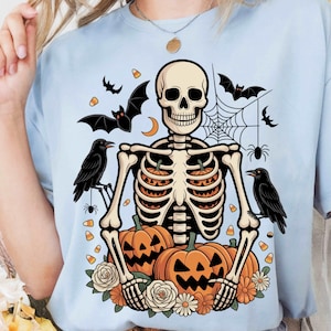 Peut inclure: T-shirt bleu clair avec un motif sur le thème d'Halloween. Le graphisme comprend un squelette, des citrouilles, des chauves-souris, des corbeaux, une toile d'araignée et des bonbons. Le squelette est entouré de citrouilles et de fleurs.
