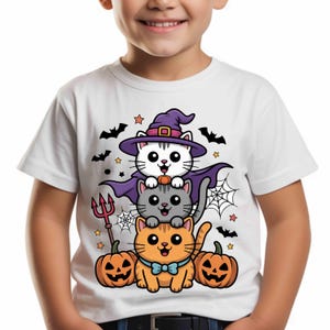 Op de afbeelding: Wit T-shirt met een Halloween-afbeelding. Het ontwerp toont drie cartoonkatten, pompoenen, vleermuizen, sterren en spinnenwebben. De bovenste kat draagt een heksenhoed en cape. De onderste kat heeft een blauwe strik.