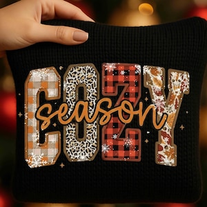 Peut inclure: Coussin noir avec l'inscription "COZY season". Les lettres sont décorées de motifs à carreaux, léopard et feuilles d'automne. Le mot "season" est en écriture cursive. Une main tient le coussin.