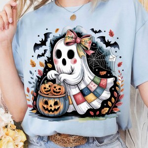 Peut inclure: T-shirt bleu clair avec un fantôme de dessin animé tenant un seau de citrouille d'Halloween avec deux citrouilles. Le fantôme porte un nœud coloré et un motif patchwork. Des chauves-souris, des feuilles et une toile d'araignée complètent le thème d'Halloween.