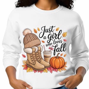 Peut inclure: Sweat-shirt blanc avec un motif sur le thème de l'automne. Le motif comprend un bonnet en tricot beige avec un pompon, des bottes beiges, une tasse de café, une citrouille et des feuilles d'automne. Le texte dit "Just a girl who loves Fall."