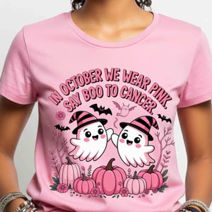 May include: Pink t-shirt with the text "IN OCTOBER WE WEAR PINK, SAY BOO TO CANCER." The shirt features a graphic of two smiling ghosts wearing witch hats, surrounded by pink pumpkins, bats, and flowers. A Halloween-themed design.