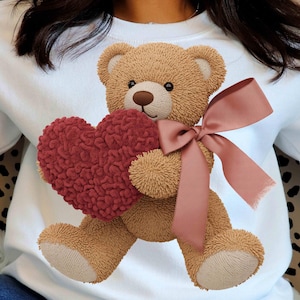 Peut inclure: Sweat-shirt blanc avec un ours en peluche tenant un cœur rouge. L'ours en peluche est brun clair avec un nez brun foncé et un nœud rose. Le cœur est de couleur rouge foncé texturé.