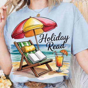 Peut inclure: T-shirt bleu clair avec un motif de scène de plage. Un transat, un livre ouvert et un cocktail sont sous un parasol rouge et jaune. Les mots "Holiday Read" sont écrits en script noir.