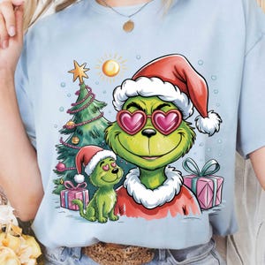 May include: Light blue t-shirt featuring a cartoon Grinch character wearing a Santa hat and heart-shaped sunglasses. A small Grinch character with a Santa hat is also present, along with a Christmas tree, gifts, and a sun.
