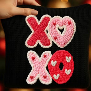 May include: Black pillow with knitted red and pink "XO" design. The "X" and "O" shapes are outlined in white and decorated with small hearts. The pillow is held by a hand.