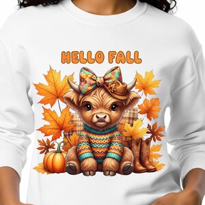 Peut inclure: Sweat-shirt blanc avec un motif de vache des Highlands portant un nœud, un chapeau et un pull, entourée de feuilles d'automne et d'une citrouille. Le texte "HELLO FALL" est au-dessus. Bottes de cowboy marron à droite.
