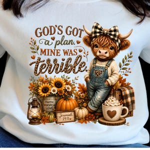 Peut inclure: Un sweat-shirt bleu clair avec une illustration de vache highland portant un nœud à carreaux et une salopette en jean. Le texte indique "GOD'S GOT a plan. MINE WAS terrible." avec des tournesols, des citrouilles, une lanterne et une tasse.