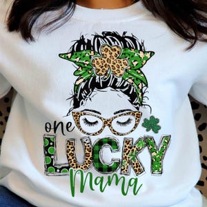 Peut inclure: Sweat-shirt blanc avec un motif graphique représentant le texte "one LUCKY Mama". Le motif comprend un visage de femme avec des lunettes, un bandeau vert et léopard et un trèfle. Les lettres sont décorées d'imprimé léopard et de carreaux verts.
