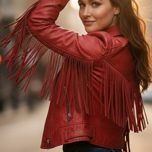 May include: Red faux leather jacket with fringe detailing on the sleeves and back. The jacket features a zipper closure and a classic biker jacket design. The model is wearing blue jeans.