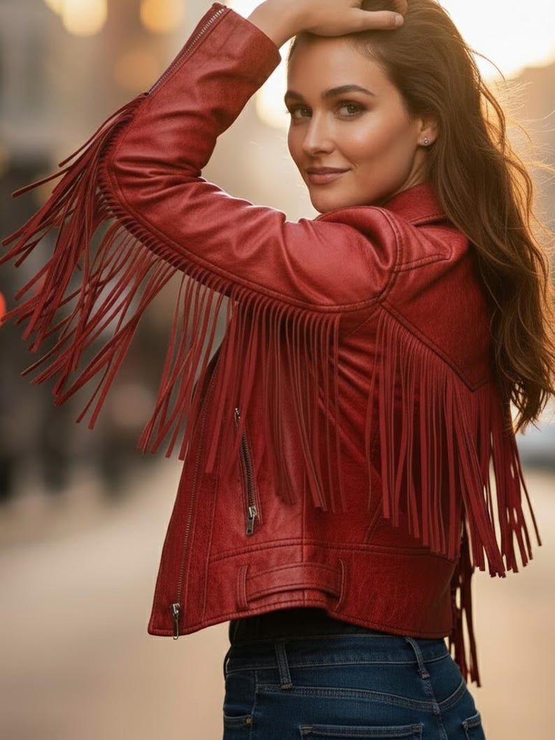 May include: Red faux leather jacket with fringe detailing on the sleeves and back. The jacket features a zipper closure and a classic biker jacket design. The model is wearing blue jeans.