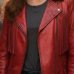 May include: A red leather jacket with fringe detailing on the chest and sleeves. The jacket has a zipper closure and a classic collar. The model is wearing a gray t-shirt and blue jeans. The jacket has a distressed look.