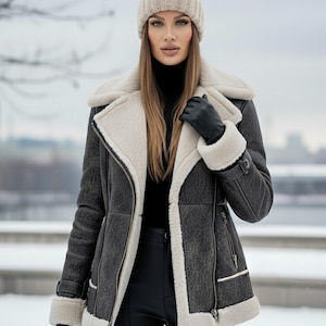 K&ouml;nnte beinhalten: Eine Frau tr&auml;gt eine dunkelgraue Shearling-Jacke mit cremefarbenem Futter und Besatz, schwarze Handschuhe, einen schwarzen Rollkragenpullover und eine beige Strickm&uuml;tze. Die Jacke hat einen Rei&szlig;verschluss und einen Schnallenriemen an den &Auml;rmeln. Der Hintergrund ist eine verschneite Szene im Freien.