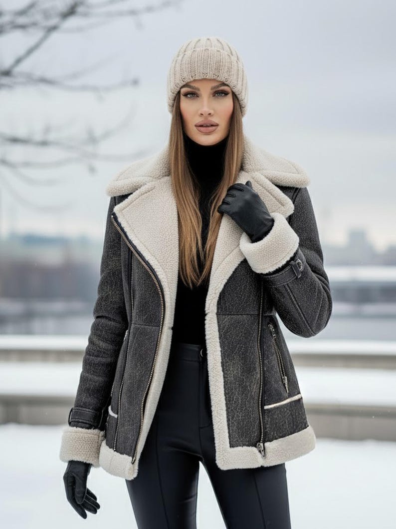 K&ouml;nnte beinhalten: Eine Frau tr&auml;gt eine dunkelgraue Shearling-Jacke mit cremefarbenem Futter und Besatz, schwarze Handschuhe, einen schwarzen Rollkragenpullover und eine beige Strickm&uuml;tze. Die Jacke hat einen Rei&szlig;verschluss und einen Schnallenriemen an den &Auml;rmeln. Der Hintergrund ist eine verschneite Szene im Freien.