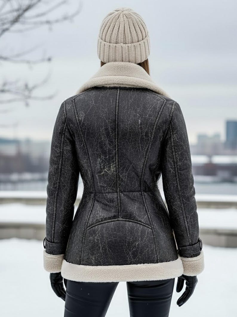 K&ouml;nnte beinhalten: Schwarze Kunstlederjacke mit cremefarbenem Shearling-Kragen und -Besatz. Die Jacke hat ein Used-Finish und wird mit einer passenden Strickm&uuml;tze und schwarzen Handschuhen getragen. Der Hintergrund zeigt eine verschneite Landschaft.