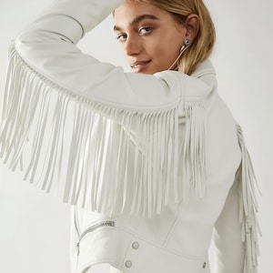 May include: White leather jacket with fringe detailing on the sleeves and back. The jacket features a zipper closure, snap buttons, and a classic biker jacket design. The fringe adds a bohemian flair to the jacket.