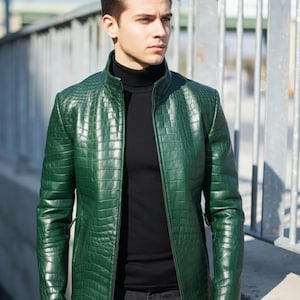 Könnte beinhalten: Grüne Lederjacke mit Krokodilhaut-Effekt. Die Jacke hat einen Stehkragen und einen Reißverschluss. Die Person trägt einen schwarzen Rollkragenpullover und dunkle Jeans.