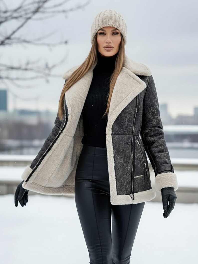 K&ouml;nnte beinhalten: Eine schwarze und graue Shearling-Jacke mit cremefarbenem Futter und Besatz. Die Jacke hat einen Rei&szlig;verschluss und wird mit schwarzen Handschuhen und einer schwarzen Hose getragen. Eine beige Strickm&uuml;tze wird ebenfalls getragen.