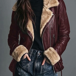 May include: A deep burgundy leather jacket featuring a wide tan faux fur collar and matching faux fur cuffs. The jacket includes silver-toned metal zippers along the front closure and side pockets, worn over a black ribbed turtleneck and blue jeans.