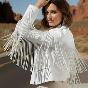 May include: A white jacket with long fringe detailing on the sleeves and back. The jacket has a zipper closure and is paired with beige trousers. The background features a desert landscape with a road.