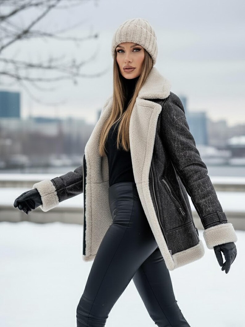 K&ouml;nnte beinhalten: Dunkelgraue Shearling-Jacke mit cremefarbenem Futter und Besatz. Die Jacke hat einen Rei&szlig;verschluss und wird mit schwarzen Handschuhen, einem schwarzen Rollkragenpullover, einer beigen Strickm&uuml;tze und schwarzen Leggings getragen. Der Hintergrund ist eine verschneite Szene im Freien.