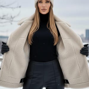 K&ouml;nnte beinhalten: Beigefarbener Shearling-Mantel, schwarzer Rollkragenpullover und schwarze Lederhose. Eine passende beige Strickm&uuml;tze wird getragen. Der Mantel ist offen und zeigt das flauschige Innenfutter. Schwarze Handschuhe vervollst&auml;ndigen den Look. Schneeiger Hintergrund.