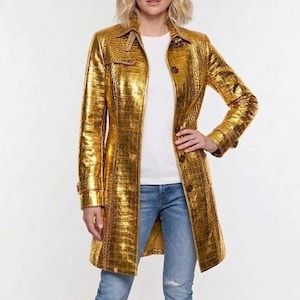 May include: A gold trench coat with a crocodile skin texture. The coat features a classic design with a collar, buttons, and long sleeves. The model is wearing a white t-shirt and blue jeans.