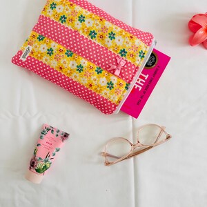 May include: A quilted fabric pouch with alternating pink polka dot and yellow floral stripes, featuring a small pink bow, a zip closure, and a white label. Included are a pink tube of hand cream, clear-framed glasses, and a pink book.