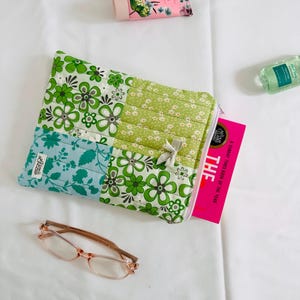 May include: A quilted fabric pouch featuring a patchwork design of green and white floral patterns, a light blue floral section, and a small grey ribbon bow. A book with a bright pink cover and the text THE is partially tucked inside the pouch.