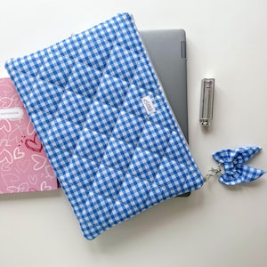 May include: A blue and white gingham laptop sleeve with a matching bow keychain. The quilted sleeve holds a laptop, next to a lipstick and a pink notebook with heart designs. The sleeve has a small white tag.