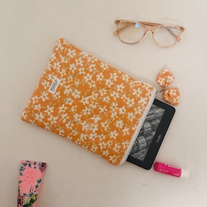 May include: An orange floral quilted Kindle sleeve with a white zip, holding a Kindle e-reader. The sleeve is decorated with small white flowers. Also pictured are glasses, lip balm, and hand cream.