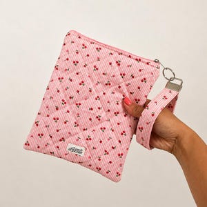 May include: A quilted rectangular zipped pouch featuring a pink and white gingham pattern with a small red cherry print. The pouch includes a matching wrist strap attached to a silver metal ring and a small white fabric label reading Lizzie.