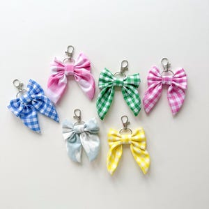 May include: Seven fabric bow keyrings in a variety of colours and patterns. The bows feature gingham checks in pink, green, and yellow, a blue and white check, and a light blue floral print. Each bow is attached to a silver-coloured metal clasp.