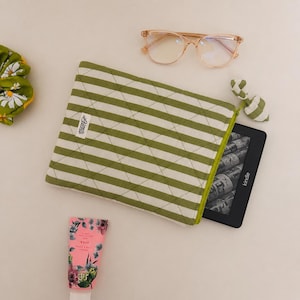 Könnte beinhalten: Eine grün-weiß gestreifte, gesteppte Kindle-Hülle mit grünem Reißverschluss und einer kleinen Stoffschlaufe. Ein Kindle E-Reader ist teilweise zu sehen. Außerdem abgebildet sind eine Brille, eine Tube Handcreme und eine grüne Blumenscrunchie.