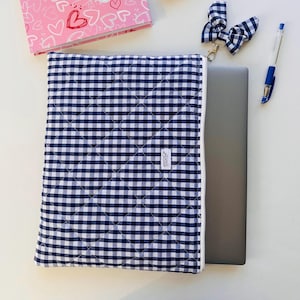Könnte beinhalten: Eine marineblaue und weiße Laptop-Hülle mit Vichy-Karo-Muster, weißem Reißverschluss und Rautensteppung. Ein passender Schlüsselanhänger mit Schleife und ein Stift liegen neben der Hülle. Ein rosa Notizbuch mit Herzen befindet sich im Hintergrund. Die Hülle hat ein kleines Etikett mit der Aufschrift "Lizzie".