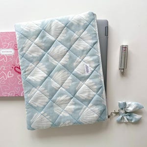 Op de afbeelding: Een lichtblauwe gewatteerde laptoptas met een wit schelpenpatroon. De hoes heeft een ritssluiting en een kleine strik-sleutelhanger. Een roze notitieboek met hartjes is zichtbaar.
