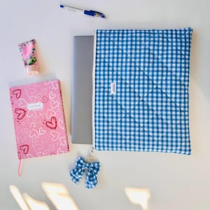 May include: A blue and white gingham laptop sleeve with a matching bow keychain. A pink notebook with heart designs and a tube of hand cream are next to the sleeve. A pen and a laptop are also in the image.