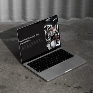 以下が含まれることがあります： フィットネスプログラムを表示するシルバーのラップトップ。画面には「Plan d'Entraînement HYROX Interm é diaire」というテキストと、運動している人の画像が表示されています。ラップトップはコンクリートの表面に開いています。