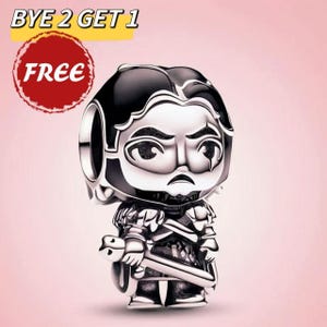 Könnte beinhalten: Ein silberfarbener Charm-Anhänger mit einer cartoonartigen Darstellung einer Figur, die ein Schwert hält. Der Anhänger hat schwarze Akzente und einen ernsten Gesichtsausdruck. Der Text "BYE 2 GET 1" und "FREE" ist im Hintergrund.
