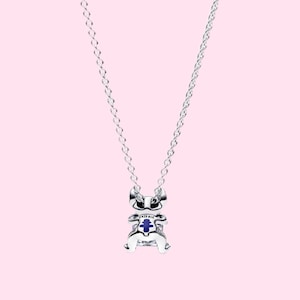 May include: A silver necklace with a charm shaped like a cartoon character. The charm has blue and white accents. The necklace hangs against a light pink background.