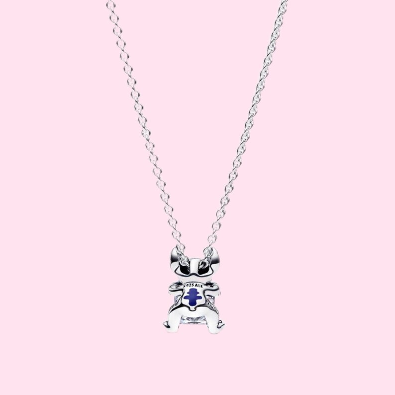 May include: A silver necklace with a charm shaped like a cartoon character. The charm has blue and white accents. The necklace hangs against a light pink background.