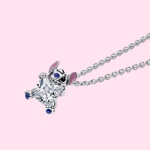 May include: A silver necklace featuring a pendant of a cartoon character with a large, square, clear gemstone body. The character has pink ears, blue eyes, and blue feet. The necklace chain is also silver, against a pink background.
