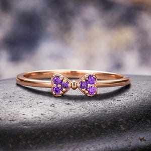 May include: A thin rose gold band ring featuring a central bow-shaped ornament. The bow is embellished with six small, round, vibrant purple gemstones, with three stones set into each side of the bow design, creating a delicate and symmetrical look.