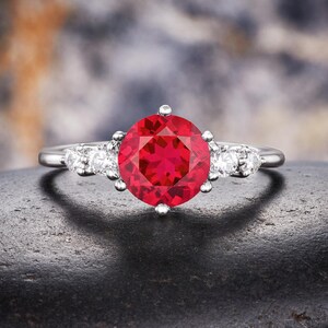 Peut inclure: Une bague argentée ornée d'une grande pierre centrale ronde d'un rouge vif maintenue par des griffes. L'anneau est agrémenté de chaque côté de trois petites pierres rondes et transparentes alignées, formant un design symétrique et poli.