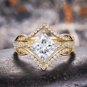 May include: A gold ring featuring a central princess-cut clear stone set in a diamond-shaped frame. The band consists of two intertwined, pavé-set gold strands that meet at the center, creating a geometric, layered appearance with small clear stones.